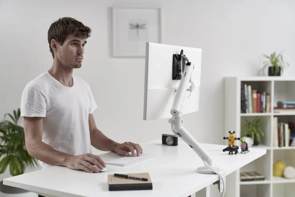 Homem trabalha em mesa com regulagem de altura e utiliza um suporte de monitor Flo branco para apoiar um monior também branco.