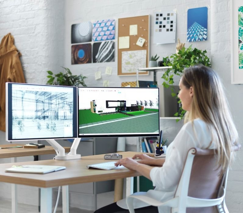 Imagem de uma mulher sentada em uma cadeira Setu Herman Miller, utilizando dois monitores apoiados em suportes Flo.