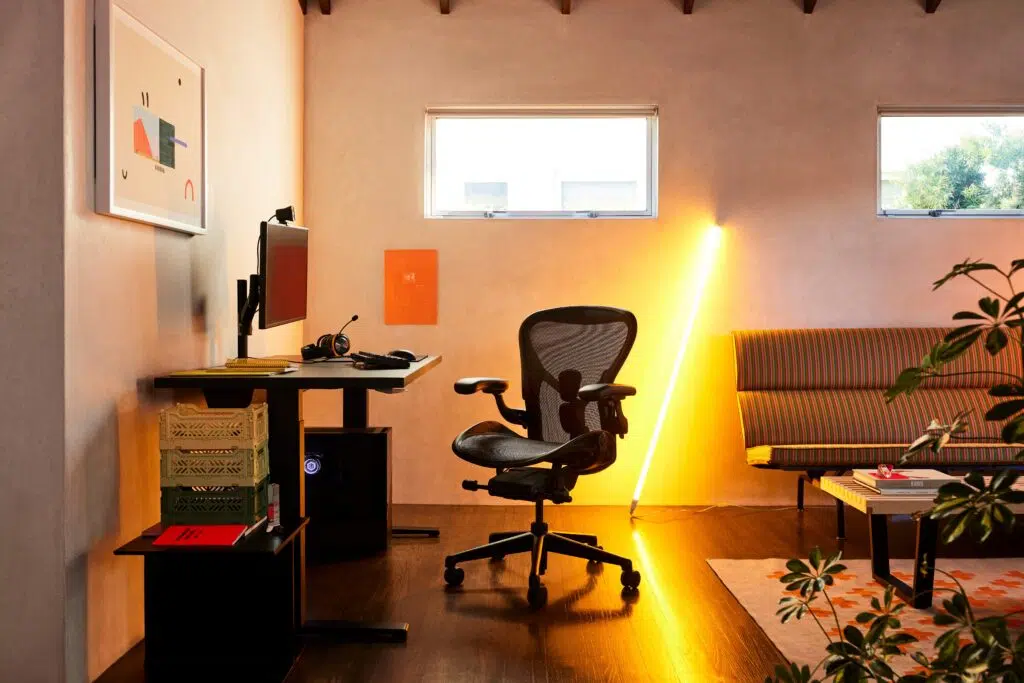 Espaço em sala de estar com cadeira Aeron, mesa Ratio e luminária na parede ao fundo.