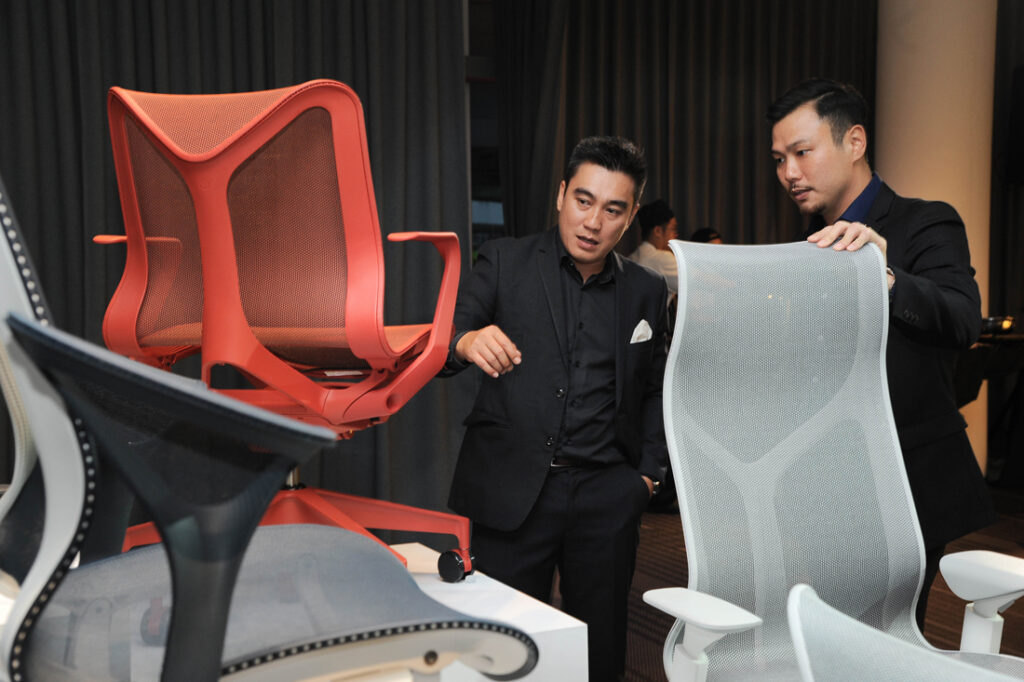 Dois homens observam, impressionados, uma cadeira Cosm Low-Back vermelha, no evento de lançamento. Um deles está com uma mão apoiada em uma cadeira Cosm High-Back na cor Mineral.