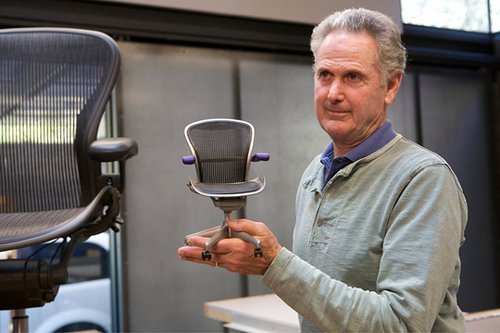 Foto de Don Chadwick segurando uma miniatura da cadeira Aeron, com o olhar dirigido para uma versão clássica da mesma, na cor Grafite.
